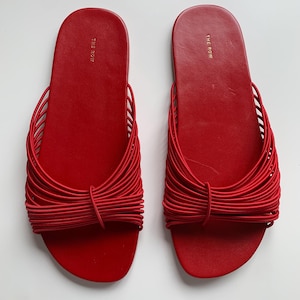 May include: A pair of bright red slide sandals featuring a flat sole and a wide strap composed of multiple thin, parallel elastic cords gathered in the center. The brand name THE ROW is printed in gold lettering on the heel of each footbed.