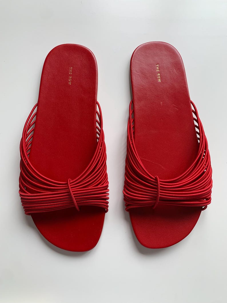 May include: A pair of bright red slide sandals featuring a flat sole and a wide strap composed of multiple thin, parallel elastic cords gathered in the center. The brand name THE ROW is printed in gold lettering on the heel of each footbed.
