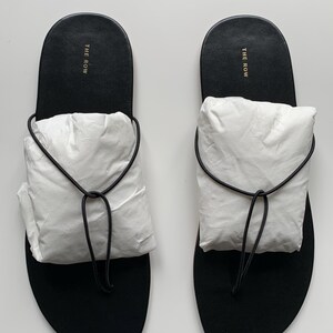 May include: A pair of black thong sandals featuring a thin black cord strap and a white paper insert covering the top of the foot area. The black footbed displays the text THE ROW in gold lettering near the heel area of each sandal.