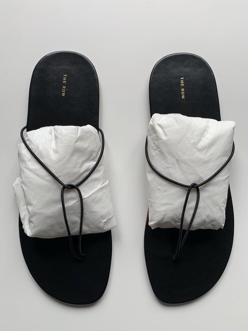 May include: A pair of black thong sandals featuring a thin black cord strap and a white paper insert covering the top of the foot area. The black footbed displays the text THE ROW in gold lettering near the heel area of each sandal.