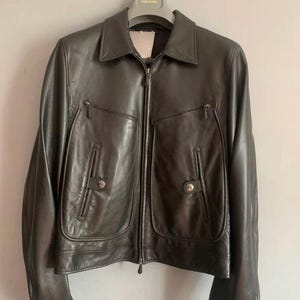 May include: Black leather jacket with a collared neckline, a front zipper, and two front pockets with button closures. The jacket has long sleeves and is hanging on a hanger.
