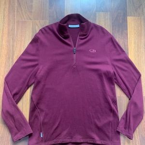 Könnte beinhalten: Ein langärmliges, weinrotes Oberteil mit einem Viertel-Reißverschluss. Das Oberteil hat einen Stehkragen und ein kleines Logo auf der Brust. Das Kleidungsstück ist aus einem weichen, wahrscheinlich sportlichen Material.