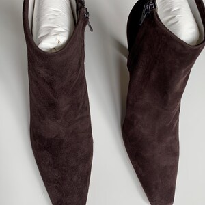 May include: A pair of dark brown ankle boots featuring a pointed toe, a side zip closure, and a block heel. The boots are crafted from a soft, matte suede-like material and contain white paper inserts to maintain their shape and structure.