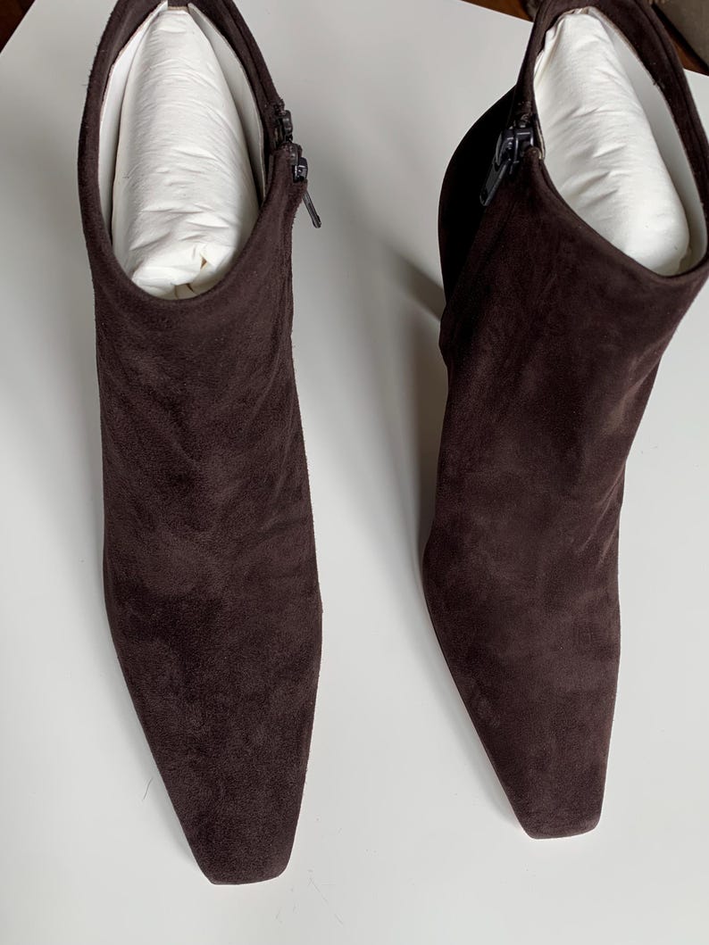 May include: A pair of dark brown ankle boots featuring a pointed toe, a side zip closure, and a block heel. The boots are crafted from a soft, matte suede-like material and contain white paper inserts to maintain their shape and structure.