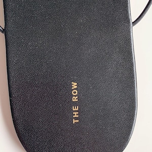 May include: A black leather sandal footbed with a rounded toe and a textured surface. The text THE ROW is printed in gold lettering on the heel area. Thin black straps extend from the sides of the sandal, which features a smooth, clean design.