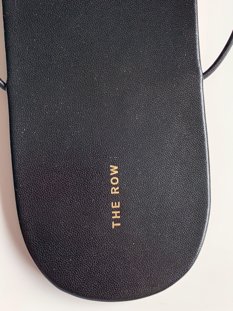 May include: A black leather sandal footbed with a rounded toe and a textured surface. The text THE ROW is printed in gold lettering on the heel area. Thin black straps extend from the sides of the sandal, which features a smooth, clean design.