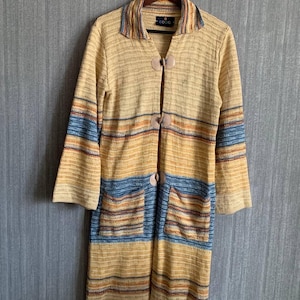 May include: A long, open-front cardigan in a beige, yellow, and blue striped pattern. The cardigan has long sleeves, two pockets, and large, round wooden buttons. The collar has the word "COOG" on it.
