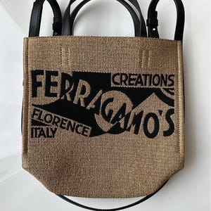 May include: A tan and black tote bag with black handles and straps. The bag features the text "FERRAGAMO'S CREATIONS FLORENCE ITALY" in a bold, stylized font. The bag has a woven texture.