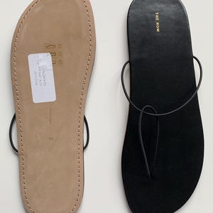 May include: A pair of flat thong sandals featuring a black suede footbed and thin black leather straps. One sandal shows the black top surface with the gold text THE ROW, while the other shows the tan leather sole with a white sticker and size 39.