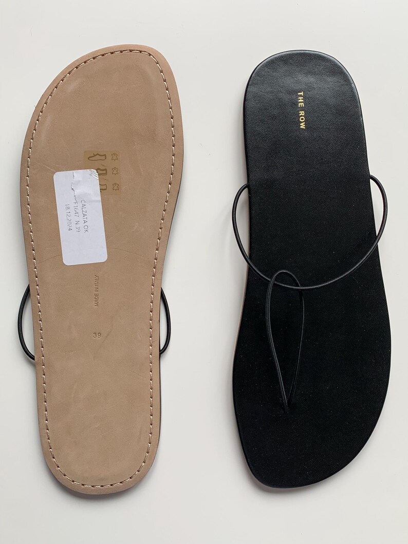 May include: A pair of flat thong sandals featuring a black suede footbed and thin black leather straps. One sandal shows the black top surface with the gold text THE ROW, while the other shows the tan leather sole with a white sticker and size 39.
