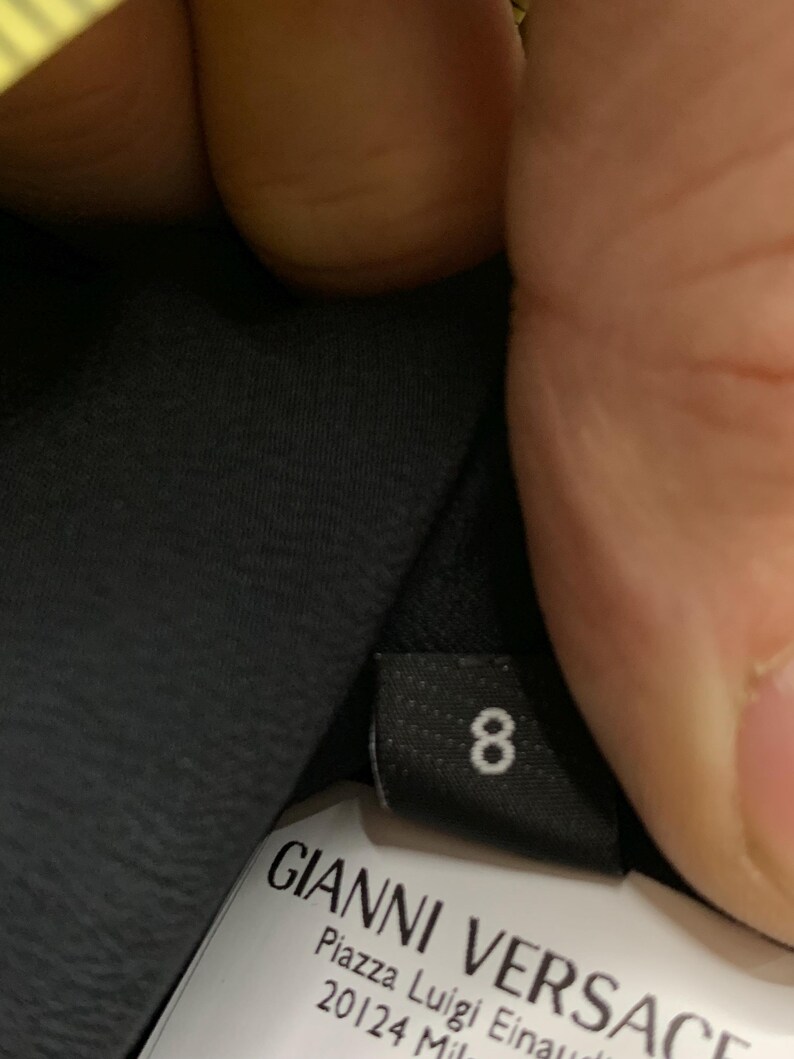 K&ouml;nnte beinhalten: Schwarzes Etikett mit der wei&szlig;en Zahl 8. Darunter ein wei&szlig;es Etikett mit dem Text "GIANNI VERSACE" und der Adresse "Piazza Luigi Einaudi 20124 Mila".