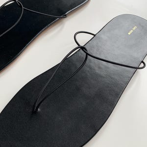 May include: A pair of black leather flat sandals featuring thin, minimalist straps that loop through the sole in a thong-style design. The footbed is smooth and black, with the brand name THE ROW printed in gold lettering near the heel area.