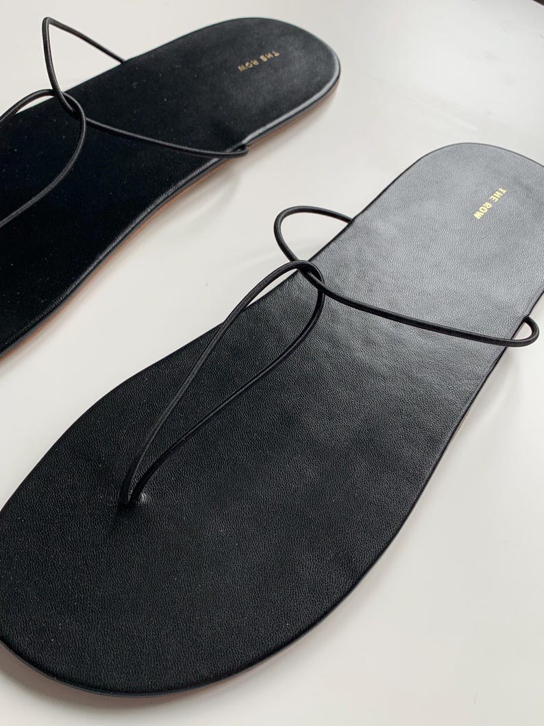 May include: A pair of black leather flat sandals featuring thin, minimalist straps that loop through the sole in a thong-style design. The footbed is smooth and black, with the brand name THE ROW printed in gold lettering near the heel area.