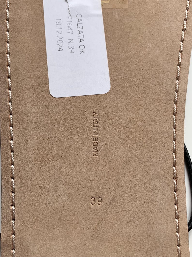 May include: The beige leather sole of a sandal features a white rectangular tag with the text CALZATA OK F1647 N.39 18.12.2024. The sole is embossed with the text MADE IN ITALY and the number 39, with light-coloured stitching along the edges.