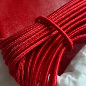 May include: A bright red sandal featuring a smooth, solid red footbed and a series of thin, parallel red elastic cords that wrap across the top. The cords are arranged in a dense, uniform pattern, creating a textured, linear appearance over the shoe.