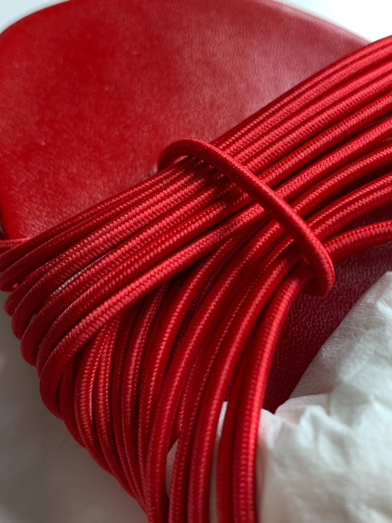 May include: A bright red sandal featuring a smooth, solid red footbed and a series of thin, parallel red elastic cords that wrap across the top. The cords are arranged in a dense, uniform pattern, creating a textured, linear appearance over the shoe.