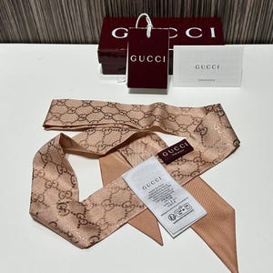 May include: A light brown silk scarf with a repeating gold-colored logo pattern. The scarf is displayed with a matching box, tag, and card, all bearing the "Gucci" brand name.