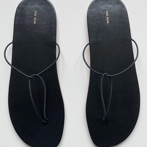 May include: A pair of black flat sandals featuring a minimalist thong-style design with thin, rounded straps that loop around the foot. The footbed is smooth and black, with the text THE ROW printed in gold lettering near the heel area of each sandal.