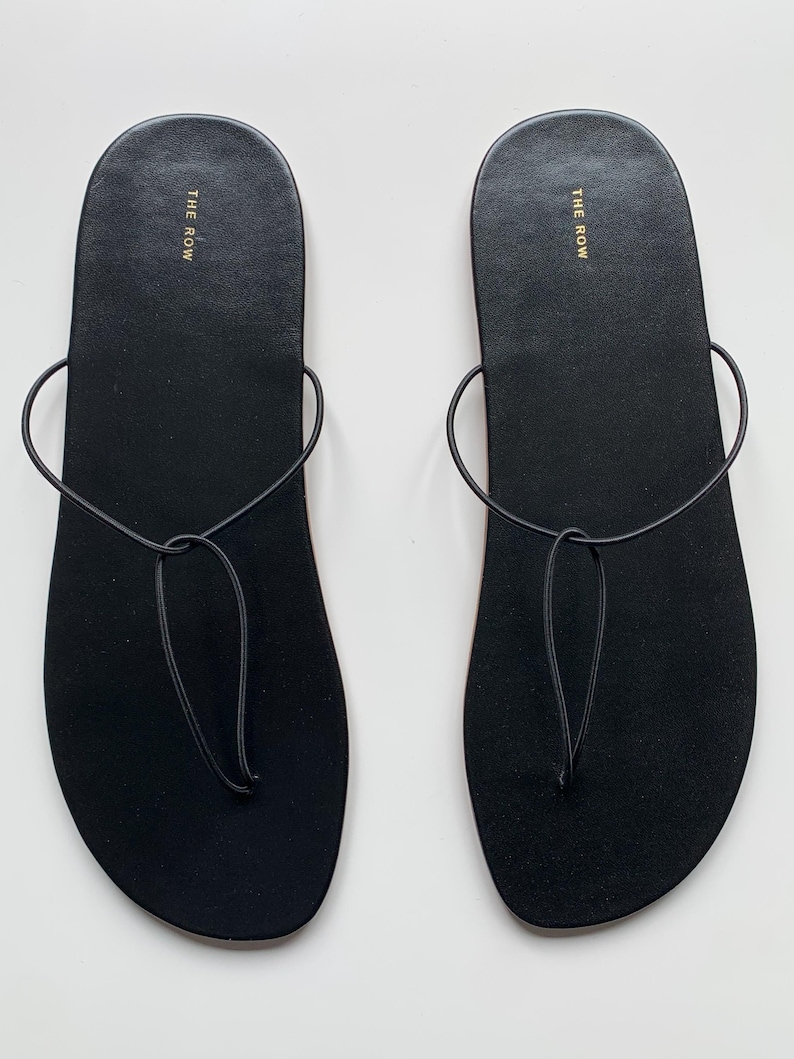 May include: A pair of black flat sandals featuring a minimalist thong-style design with thin, rounded straps that loop around the foot. The footbed is smooth and black, with the text THE ROW printed in gold lettering near the heel area of each sandal.