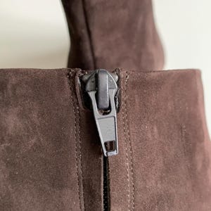 May include: A dark brown ankle boot featuring a soft suede-like texture and a vertical zip closure on the back. The zip pull is a matte dark grey metal, positioned at the top of the shaft, showing detailed stitching along the seam.