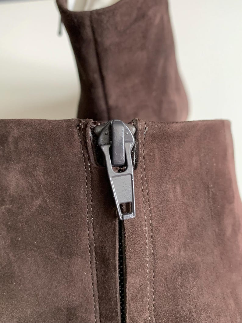 May include: A dark brown ankle boot featuring a soft suede-like texture and a vertical zip closure on the back. The zip pull is a matte dark grey metal, positioned at the top of the shaft, showing detailed stitching along the seam.