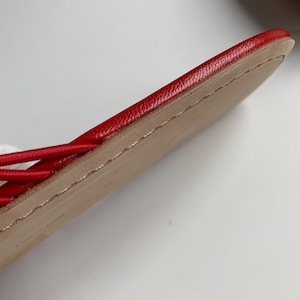 May include: A red sandal featuring a smooth, vibrant red leather upper strap and a flat, tan-colored sole. The edge of the sole shows visible stitching detail along the perimeter, connecting the upper material to the base of the footwear.