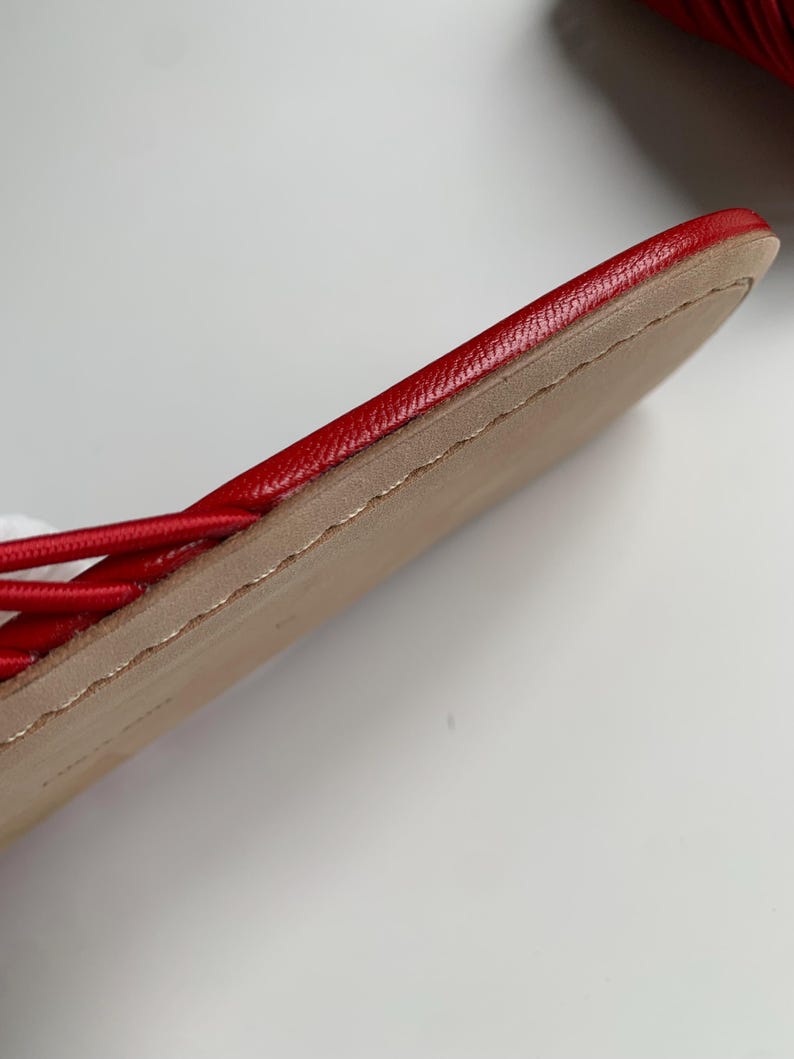 May include: A red sandal featuring a smooth, vibrant red leather upper strap and a flat, tan-colored sole. The edge of the sole shows visible stitching detail along the perimeter, connecting the upper material to the base of the footwear.