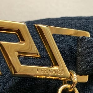 K&ouml;nnte beinhalten: Nahaufnahme eines goldfarbenen Metallaccessoires mit dem eingravierten Markennamen "VERSACE". Das Accessoire hat ein geometrisches Design mit Kettenbefestigung und ist vor einem dunkelblauen Stoffhintergrund platziert.
