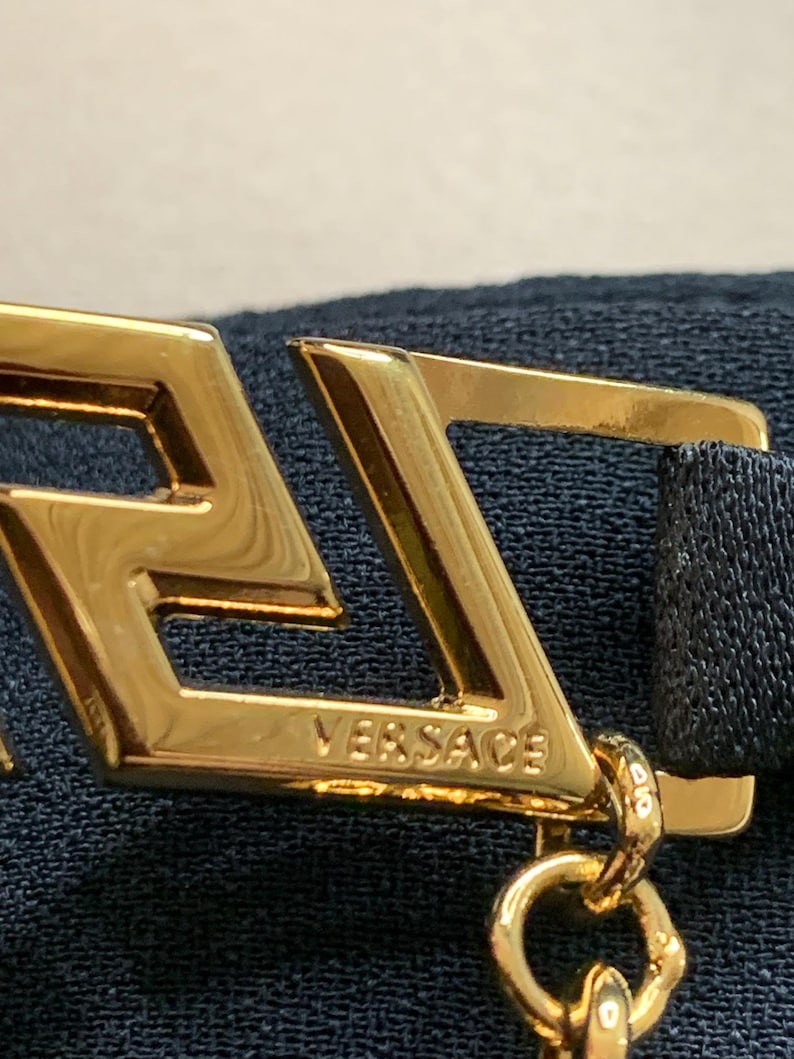 K&ouml;nnte beinhalten: Nahaufnahme eines goldfarbenen Metallaccessoires mit dem eingravierten Markennamen "VERSACE". Das Accessoire hat ein geometrisches Design mit Kettenbefestigung und ist vor einem dunkelblauen Stoffhintergrund platziert.