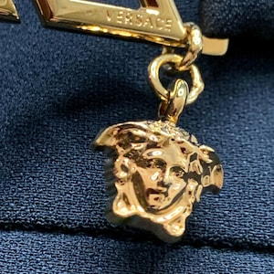 K&ouml;nnte beinhalten: Nahaufnahme eines goldfarbenen Versace-Anh&auml;ngers. Der Anh&auml;nger zeigt das Versace-Logo und einen Medusa-Kopf-Anh&auml;nger. Der Schmuck befindet sich vor einem dunkelblauen Stoffhintergrund.