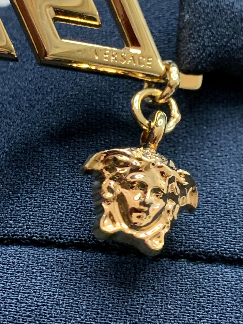 K&ouml;nnte beinhalten: Nahaufnahme eines goldfarbenen Versace-Anh&auml;ngers. Der Anh&auml;nger zeigt das Versace-Logo und einen Medusa-Kopf-Anh&auml;nger. Der Schmuck befindet sich vor einem dunkelblauen Stoffhintergrund.