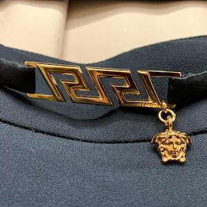K&ouml;nnte beinhalten: Ein schwarzes Choker-Halsband mit einem goldfarbenen rechteckigen Verschluss mit griechischem Schl&uuml;sseldesign. Ein goldener Medusa-Kopf-Anh&auml;nger baumelt am Verschluss. Das Halsband befindet sich vor einem dunkelblauen Stoffhintergrund.