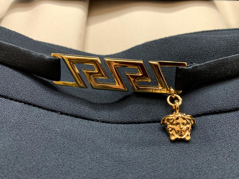 K&ouml;nnte beinhalten: Ein schwarzes Choker-Halsband mit einem goldfarbenen rechteckigen Verschluss mit griechischem Schl&uuml;sseldesign. Ein goldener Medusa-Kopf-Anh&auml;nger baumelt am Verschluss. Das Halsband befindet sich vor einem dunkelblauen Stoffhintergrund.