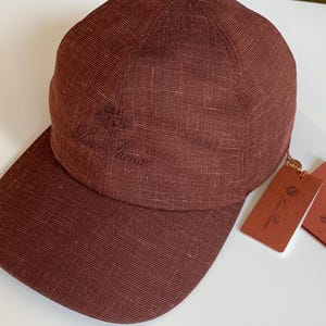 Könnte beinhalten: Eine weinrote Baseballkappe mit gebogenem Schirm. Die Kappe hat das gestickte Logo "Loro Piana" auf der Vorderseite und enthält angebrachte Etiketten. Die Kappe ist aus einem strukturierten Stoff.