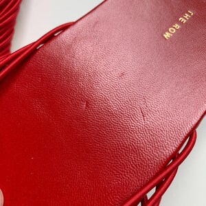 May include: A bright red slide sandal featuring a textured footbed with the text THE ROW printed in gold lettering. The sandal has a series of thin, rounded red straps extending from the sides of the sole, creating a cage-like design.