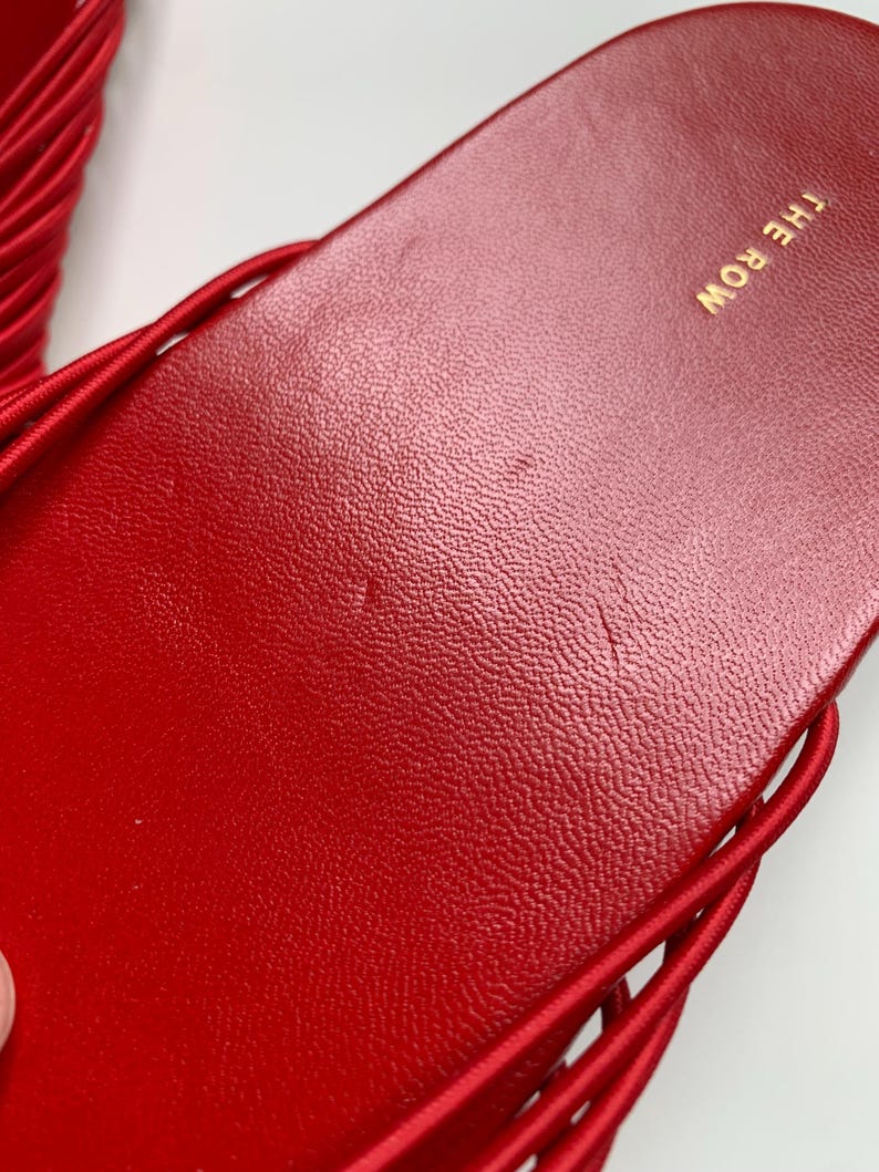 May include: A bright red slide sandal featuring a textured footbed with the text THE ROW printed in gold lettering. The sandal has a series of thin, rounded red straps extending from the sides of the sole, creating a cage-like design.