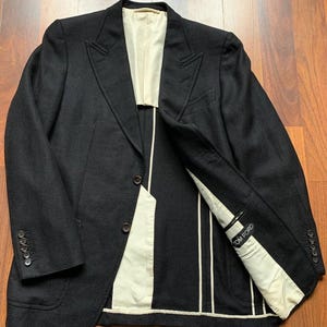 May include: A dark navy blazer with a textured weave. The jacket features a notched lapel, two-button closure, and multiple pockets. The interior lining is a cream color with the brand name "TOM FORD" visible.