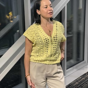Pode incluir: Um top de crochê amarelo claro com decote em V e mangas curtas. O top é combinado com calças bege. O design de crochê apresenta um padrão aberto e arejado. A roupa é adequada para uso casual.