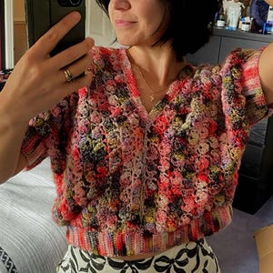 May include: A woman wearing a multi-coloured crochet top with a v-neck and short sleeves. The top has a textured, bobbly design. She is also wearing black and white trousers with a bow pattern.