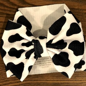 May include: A white headband with a black and white cow print bow. The headband has a care label that reads "ic & imported re wash COLD with flat to dry or tumble d Do not bleach Ratch"