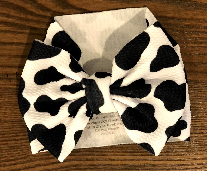 May include: A white headband with a black and white cow print bow. The headband has a care label that reads "ic & imported re wash COLD with flat to dry or tumble d Do not bleach Ratch"