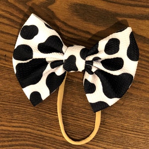 May include: A black and white cow print hair bow with a nude elastic headband.