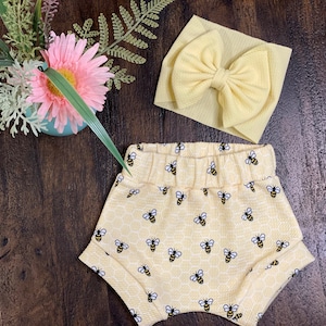 May include: Yellow baby shorts with a honeycomb pattern and black bees. A yellow bow headband is also included.
