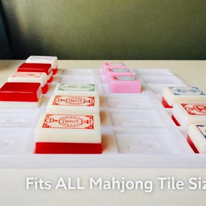 May include: A white Mahjong tile tray with tiles in red, pink, and white. The tiles are arranged in rows. The text "Fits ALL Mahjong Tile Sizes" is at the bottom of the image.