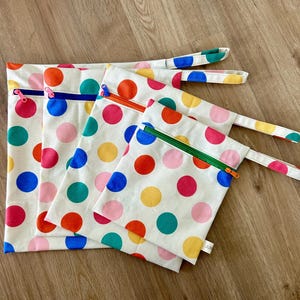 Könnte beinhalten: Vier wiederverwendbare Reißverschlusstaschen mit bunten Punkten auf weißem Hintergrund. Die Taschen sind aus Stoff und haben einen Reißverschluss. Die Taschen sind übereinander gestapelt, wobei die größte Tasche unten und die kleinste Tasche oben liegt. Die Taschen sind alle unterschiedlich groß.
