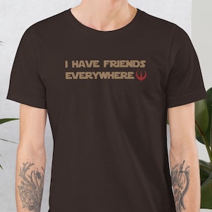 Può includere: Una maglietta marrone con il testo "I have friends everywhere" e un simbolo rosso.