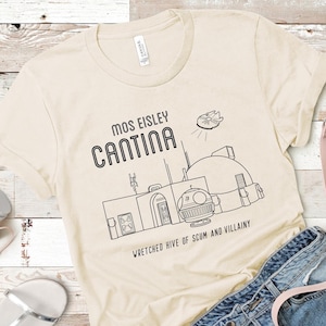 Könnte beinhalten: Ein beiges T-Shirt mit einer schwarzen Strichzeichnung der Mos Eisley Cantina aus Star Wars. Der Text "Mos Eisley Cantina" befindet sich über der Zeichnung und der Text "Wretched hive of scum and villainy" unter der Zeichnung.