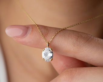 May include: A gold-colored necklace with a large, oval-shaped, clear gemstone pendant. The pendant is held in place by four prongs. The necklace chain is delicate and thin. The background is blurred.