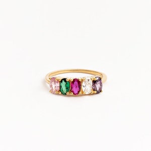 Op de afbeelding: Een gouden ring met vijf ovaal geslepen edelstenen op een rij. De stenen zijn gezet in een klauwzetting en vertonen een verscheidenheid aan kleuren, waaronder lichtroze, levendig groen, diep magenta, helder wit en donkerpaars.