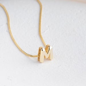 May include: A gold-colored necklace featuring a pendant in the shape of the letter "M". The pendant is a bold, block letter design. The chain is a delicate, linked style. The necklace is displayed against a white background.